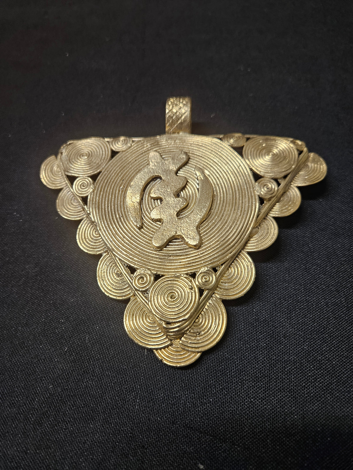 Oversized Gye Nyame Adinkra brass pendant from Ghana with triangular form, central symbol, concentric rings, and spiral decoration.